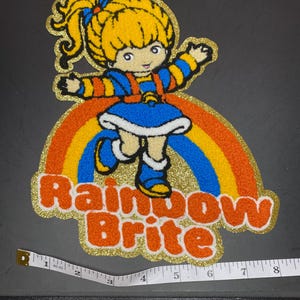 May include: A Rainbow Brite patch with a gold glitter border, featuring the character with yellow hair, a blue dress, and a rainbow. The text Rainbow Brite is in orange. A white measuring tape shows the patch is approximately 8 inches wide.