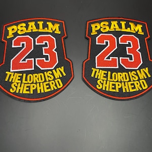 May include: Two shield-shaped embroidered patches with black backgrounds and red borders. Each patch features the yellow text PSALM 23 at the top, large red numbers 23 in the center, and the yellow text THE LORD IS MY SHEPHERD at the bottom.