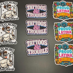 May include: Three sets of stickers featuring three distinct designs: a black and white illustration of a person with the text 'GOOD MOMS Say Bad Words', a black sticker with the text 'TATTOOS & TROUBLE', and a colorful floral design with 'PROUD LATINA'.