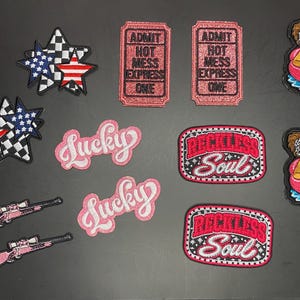 May include: A collection of embroidered patches including two star clusters with American flag and checkered patterns, two pink ticket patches reading ADMIT HOT MESS EXPRESS ONE, two pink Lucky script patches, two pink rifle patches, and two flamingo float patches.
