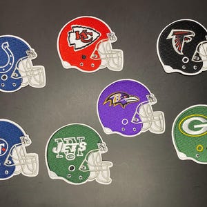 May include: A collection of seven embroidered patches shaped like American football helmets. The patches feature various team logos and colors, including blue, red, black, purple, and green, each with a white face mask design stitched on.