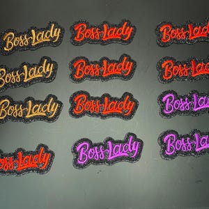 May include: A collection of eleven embroidered patches featuring the text Boss lady in cursive script. The patches have a black glittery border and are available in gold, red, and purple thread colors, arranged in a grid on a dark surface.