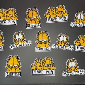May include: A collection of twelve embroidered patches featuring the orange tabby cat Garfield in various poses. Some patches show the character with the text HAVE FUN or GARFIELD in black lettering on a white background, while others show the name.