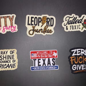 Può includere: Sei toppe ricamate con vari disegni e testi, tra cui PETTY & not sorry, LEOPARD JUNKIE con un fulmine, Tatted & TOXIC con una rosa, I'M A RAY OF SUNSHINE WITH A CHANCE OF HURRICANE, WELCOME TO TEXAS e ZERO FUCKS GIVEN!