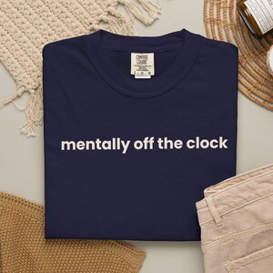 Puede incluir: Una camiseta de cuello redondo azul marino con el texto blanco "mentally off the clock" impreso en el pecho. La camiseta tiene una etiqueta de la marca Comfort Colors en el cuello interior que indica la talla grande, hecha de algodón.