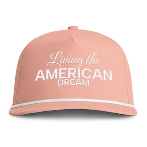 Puede incluir: Una gorra de béisbol color melocotón con el texto bordado en blanco Living the AMERICAN DREAM en el panel frontal. La gorra incluye un cordón blanco decorativo que recorre horizontalmente la base de la corona, sobre la visera curva.
