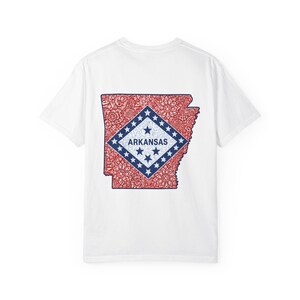 May include: A white short-sleeve t-shirt featuring a graphic on the back of the state of Arkansas. The state outline is filled with a red and white floral mandala pattern, centered with a blue diamond containing the word ARKANSAS and stars.