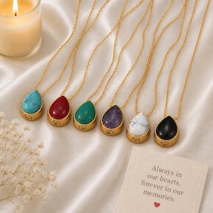 May include: A collection of teardrop-shaped pendant necklaces in various colors, including turquoise, red, green, purple, white, and black. Each pendant is set in a gold-colored bezel and hangs from a delicate gold chain. A card reads "Always in our hearts, forever in our memories."