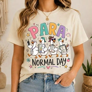 May include: A cream-colored t-shirt featuring a graphic of four cartoon ducks dressed as school staff. The text above reads PARA and below reads NORMAL DAY. The ducks hold items like a walkie-talkie, a schedule, a clipboard, and a pencil.