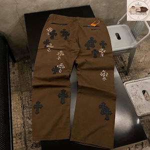 May include: Brown pants with multiple cross designs in black and leopard print. The pants have a relaxed fit and a tag attached to the back pocket. The crosses are embroidered on the front and back of the pants.