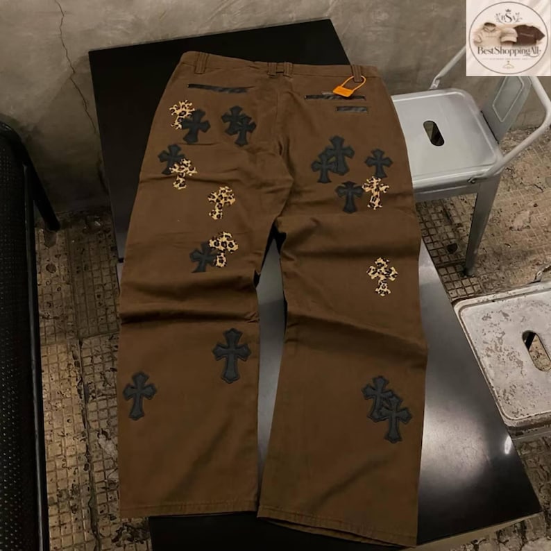 May include: Brown pants with multiple cross designs in black and leopard print. The pants have a relaxed fit and a tag attached to the back pocket. The crosses are embroidered on the front and back of the pants.
