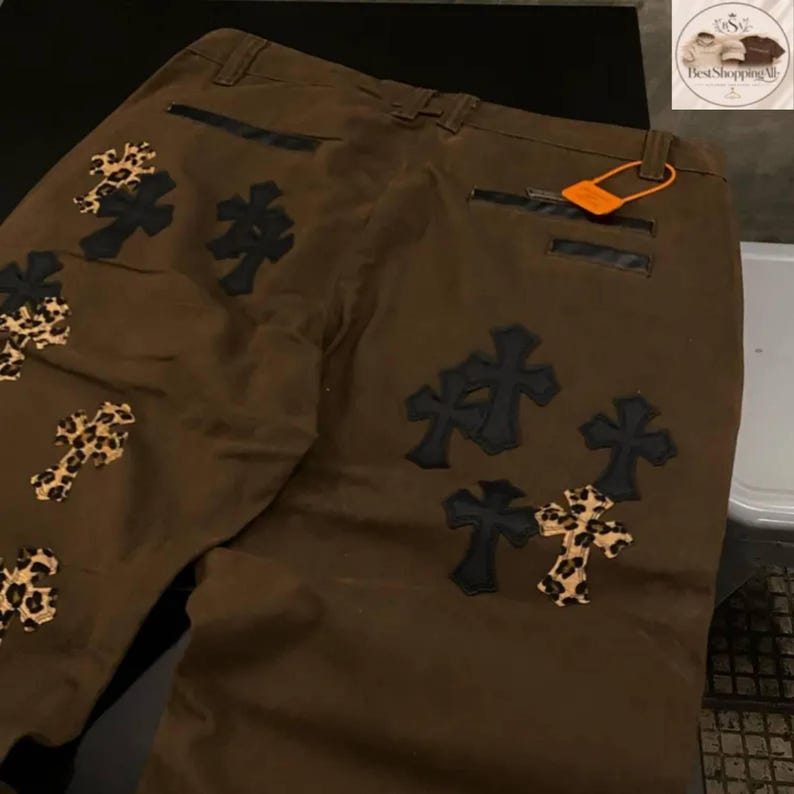 May include: Brown pants with multiple cross patches. Some crosses are black, others feature a leopard print design. The pants have a classic five-pocket design and a small orange tag.