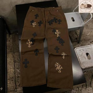 May include: Brown denim pants with multiple cross designs. Some crosses are solid black, while others feature a leopard print pattern. The pants have a classic five-pocket design and a straight leg fit.