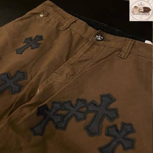 May include: Brown pants with multiple black cross appliques. The pants have a button closure and belt loops. The crosses are stitched onto the fabric. The pants are made of a sturdy material.