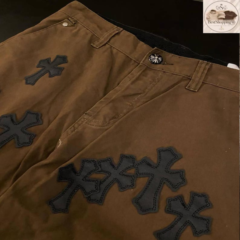May include: Brown pants with multiple black cross appliques. The pants have a button closure and belt loops. The crosses are stitched onto the fabric. The pants are made of a sturdy material.