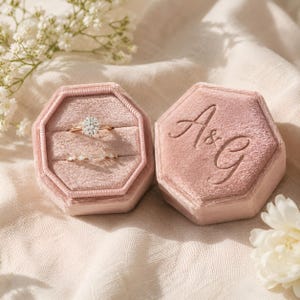 May include: Two pink velvet hexagonal ring boxes, one open with a diamond ring and a smaller ring, the other closed with the initials "A&G". They rest on a cream-coloured surface.