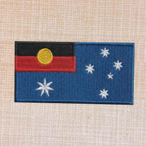 May include: A rectangular embroidered patch featuring a blue background with a black and red horizontal stripe section in the upper left corner. A yellow circle is centered in the black and red area, and five white stars are arranged on the blue field.