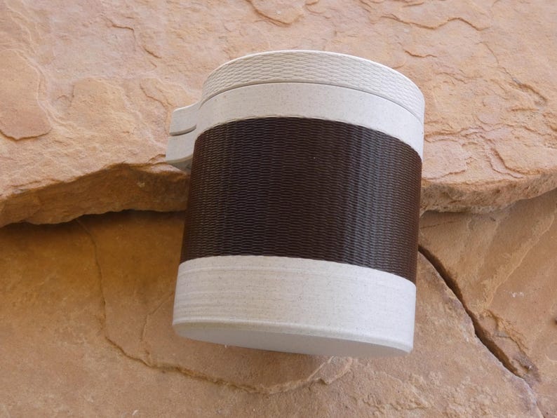 Op de afbeelding: Een cilindrische reismok met een getextureerde bruine band en witte boven- en onderdelen. De mok heeft een handvat en is ontworpen om dranken warm of koud te houden. De mok staat op een rotsachtig oppervlak.