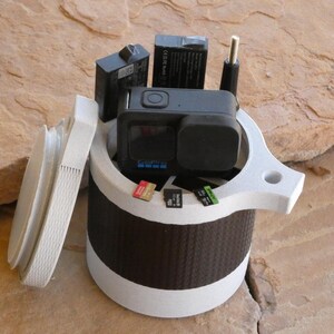 Op de afbeelding: Een witte en zwarte GoPro camerakoffer met een deksel, met een zwarte action camera, batterijen en geheugenkaarten. De koffer heeft een cilindrisch ontwerp met een getextureerde zwarte band en een witte boven- en onderkant.