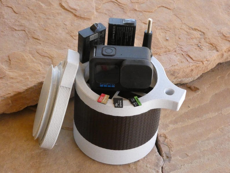 Op de afbeelding: Een witte en zwarte GoPro camerakoffer met een deksel, met een zwarte action camera, batterijen en geheugenkaarten. De koffer heeft een cilindrisch ontwerp met een getextureerde zwarte band en een witte boven- en onderkant.
