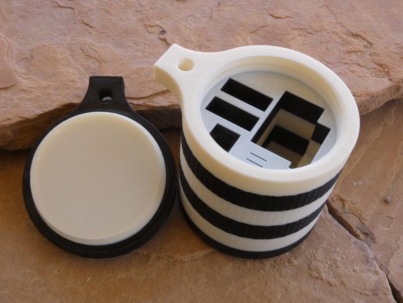 Puede incluir: Un conjunto de contenedor y tapa impresos en 3D. El contenedor es blanco con rayas horizontales negras y un interior abierto. La tapa es negra con un inserto circular blanco. El conjunto est&aacute; dise&ntilde;ado para el almacenamiento.