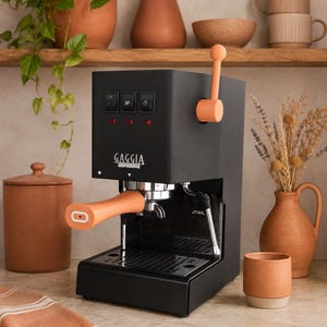 May include: A matte black Gaggia espresso machine featuring a peach-colored portafilter handle and a matching peach-colored steam wand lever. The front panel includes three black rectangular power and function switches with red indicator lights.