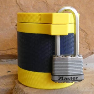 Op de afbeelding: Een gele en marineblauwe container met een zilveren hangslot. De container heeft een textuurontwerp en is beveiligd met een Master Lock-hangslot. De container is cilindrisch met gele banden aan de boven- en onderkant.