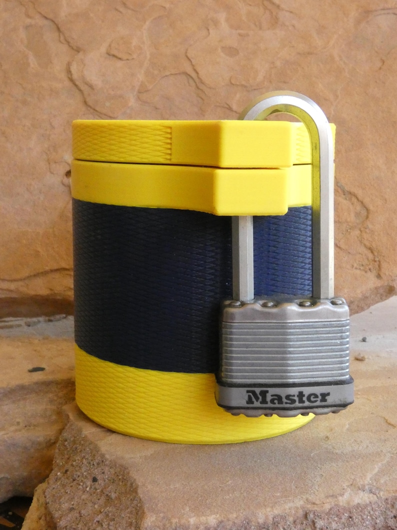 Op de afbeelding: Een gele en marineblauwe container met een zilveren hangslot. De container heeft een textuurontwerp en is beveiligd met een Master Lock-hangslot. De container is cilindrisch met gele banden aan de boven- en onderkant.