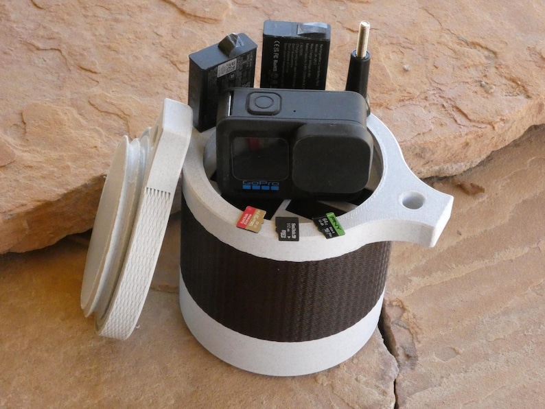 Op de afbeelding: Een witte en bruine GoPro camerakoffer met een deksel, met een zwarte GoPro camera, batterijen en geheugenkaarten. De koffer heeft een handvat en een gestreepte bruine band in het midden.