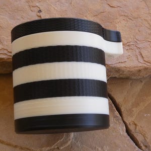 Puede incluir: Una taza cil&iacute;ndrica con asa, con rayas horizontales alternas en blanco y negro. La taza est&aacute; hecha de un material texturizado y se apoya sobre una superficie de piedra natural. El asa es blanca.