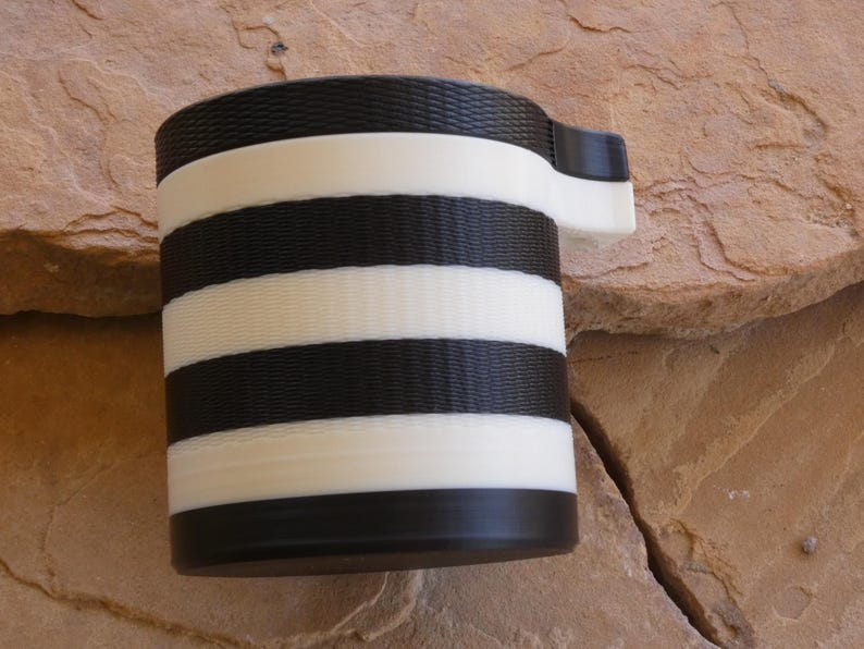 Puede incluir: Una taza cil&iacute;ndrica con asa, con rayas horizontales alternas en blanco y negro. La taza est&aacute; hecha de un material texturizado y se apoya sobre una superficie de piedra natural. El asa es blanca.