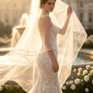 Puede incluir: Un vestido de novia color marfil hasta el suelo con intrincados bordados de encaje blanco, delicados adornos de perlas y tirantes finos. El vestido se combina con un velo largo, transparente y fluido adornado con pequeñas perlas.