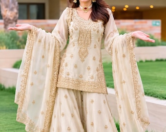 Peut inclure: Un ensemble trois pièces couleur crème composé d'une tunique à manches longues et d'un pantalon large, tous deux ornés de broderies dorées complexes. Un dupatta transparent assorti à bordure dorée est drapé sur les bras.