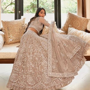 Puede incluir: Un conjunto de lehenga choli beige con intrincados bordados plateados, lentejuelas y espejos en la falda acampanada y la blusa de manga larga. El conjunto incluye un dupatta transparente a juego con un borde decorativo y adornos detallados.