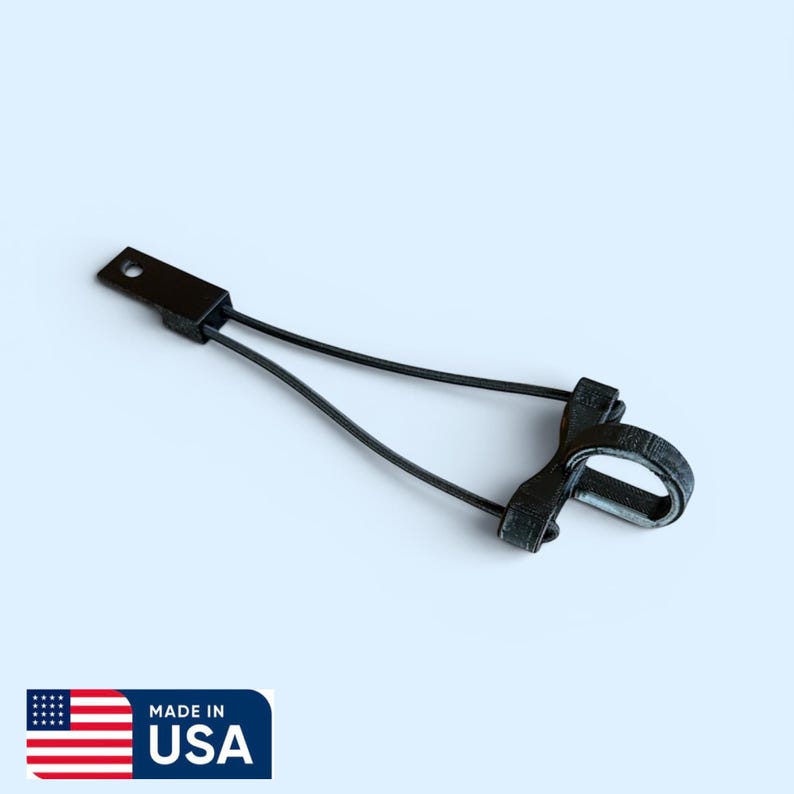 May include: A black, 3D-printed tethering device featuring a flat rectangular mounting tab with a circular hole, connected by two thin, flexible black cords to a small, circular ring component designed for securing items in place.