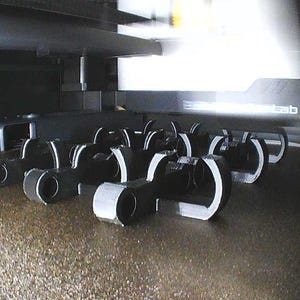 May include: A collection of black, C-shaped plastic clips arranged in rows on the textured build plate of a 3D printer. The objects feature a circular opening at one end and a curved, open-ended structure, captured from an elevated angle.