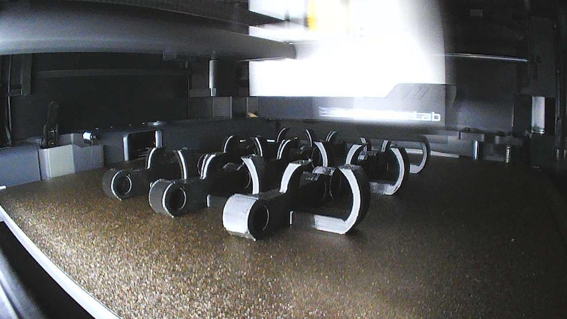 May include: A collection of black, C-shaped plastic clips arranged in rows on the textured build plate of a 3D printer. The objects feature a circular opening at one end and a curved, open-ended structure, captured from an elevated angle.