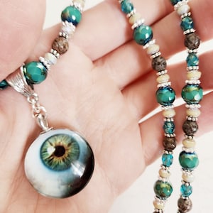 May include: A silver chain necklace with a large, round, blue and green eye pendant. The necklace is made with brown, cream, and blue beads.