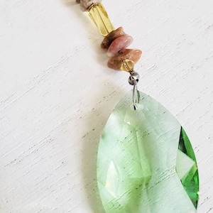 May include: A decorative pendant featuring a faceted green crystal teardrop. The pendant is adorned with a strand of small, colorful beads, including yellow, brown, and pink stones. The pendant hangs from a metal clasp.