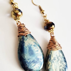 May include: Dangle earrings with gold-coloured hooks and accents. Each earring has a teardrop-shaped, blue and white marbled stone wrapped with copper wire. A faceted, dark gold bead sits above the wire-wrapped stone.