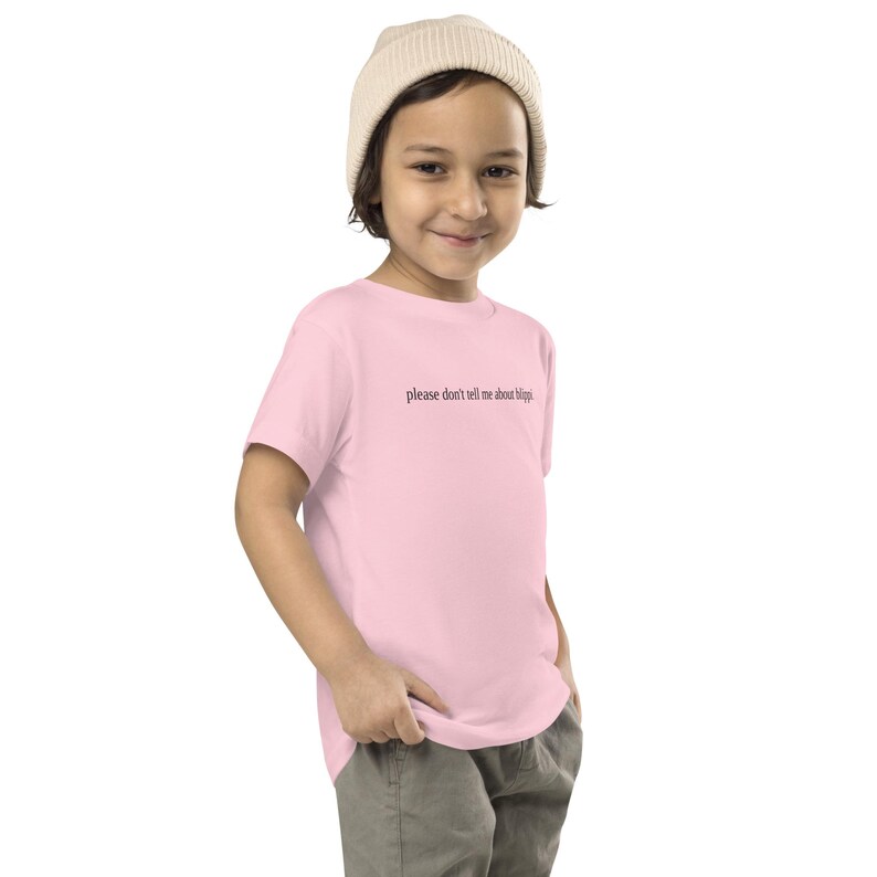 May include: A light pink toddler t-shirt with the text "please don't tell me about hippies" in black. The child is wearing a cream-colored beanie and gray pants.