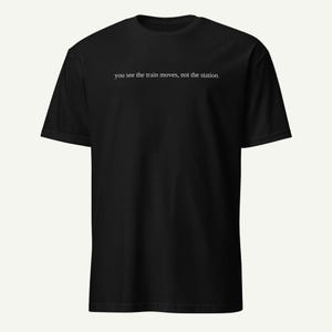 May include: Black t-shirt with the text "you see the train moves, not the station." The shirt has short sleeves and a crew neck. The text is in a simple, sans-serif font.
