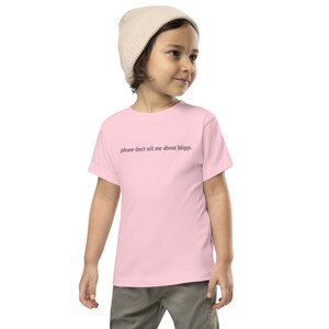 May include: A light pink t-shirt with the text "please don't tell me about blippi." The shirt is worn by a child, who is also wearing a beige beanie and gray pants.
