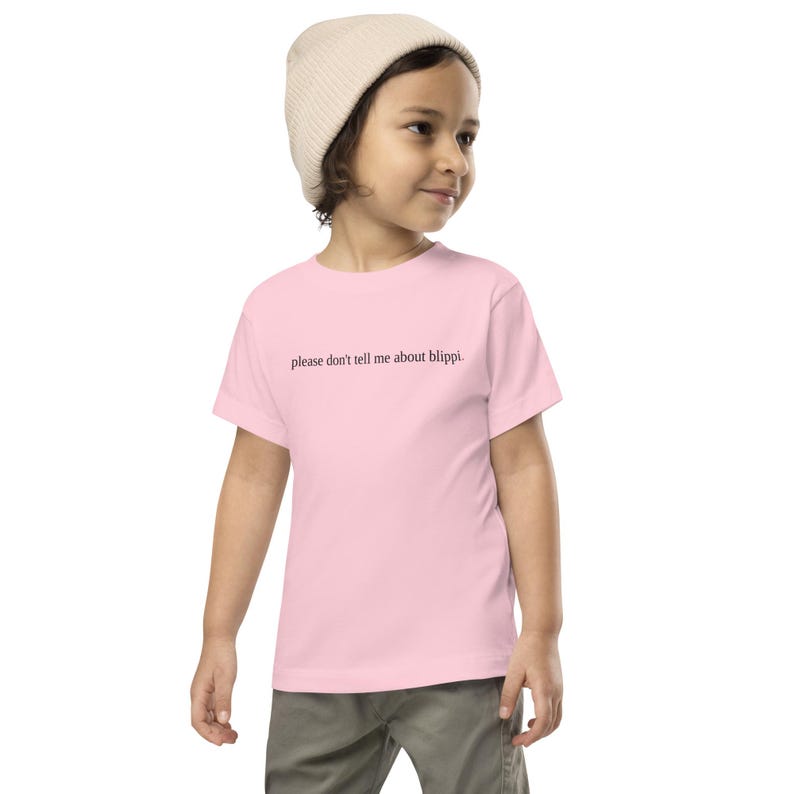 May include: A light pink t-shirt with the text "please don't tell me about blippi." The shirt is worn by a child, who is also wearing a beige beanie and gray pants.