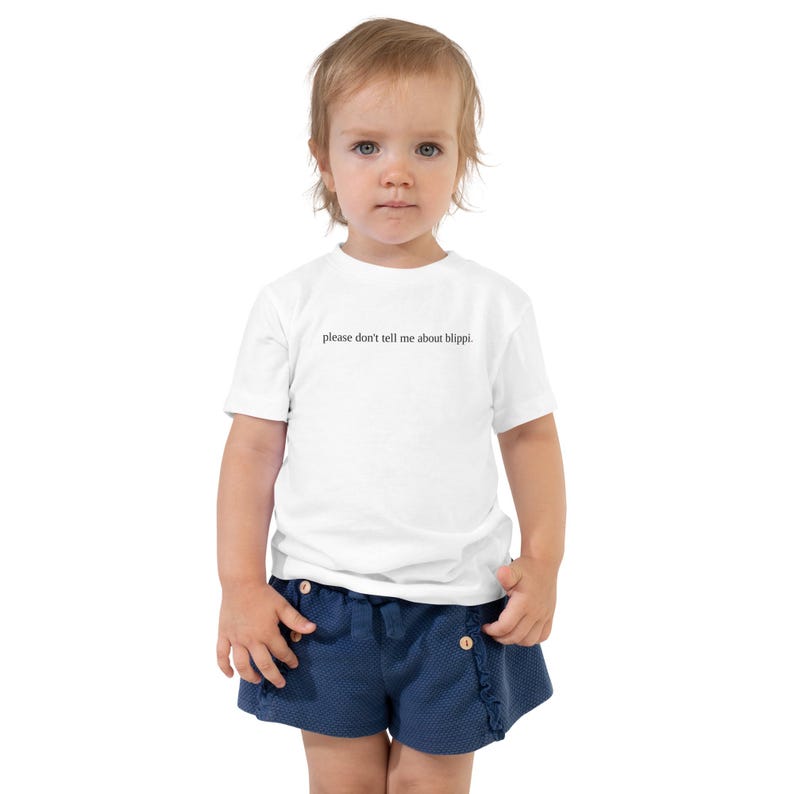 May include: A toddler wearing a white t-shirt with the text "please don't tell me about blippi." The child is also wearing navy blue shorts.