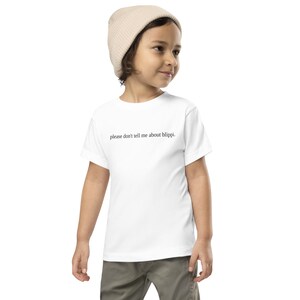 May include: A white toddler t-shirt with the text "please don't tell me about blippi." The shirt is worn by a child wearing a beige beanie and khaki pants.