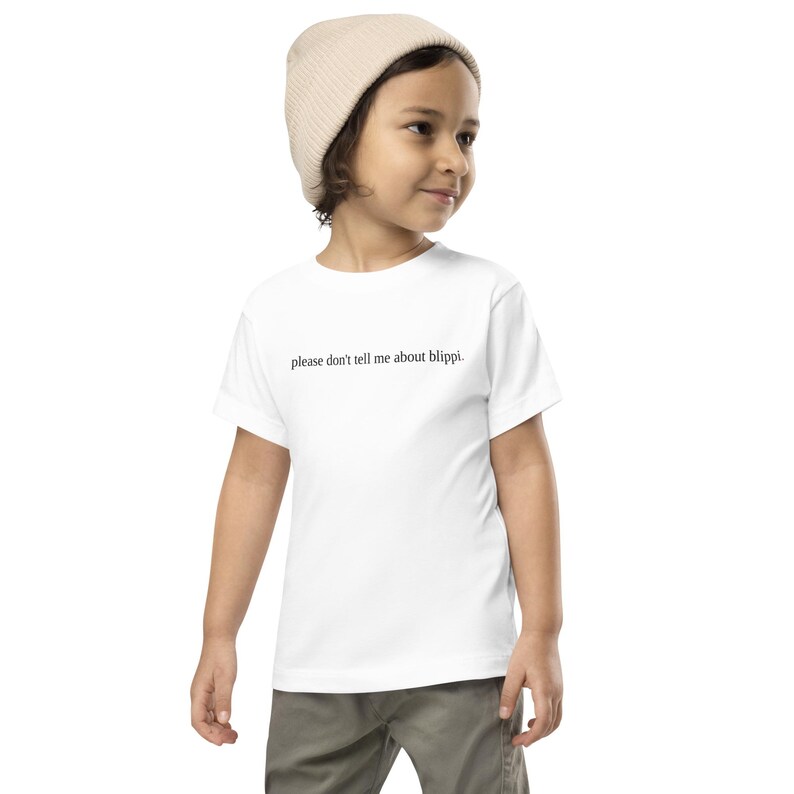 May include: A white toddler t-shirt with the text "please don't tell me about blippi." The shirt is worn by a child wearing a beige beanie and khaki pants.