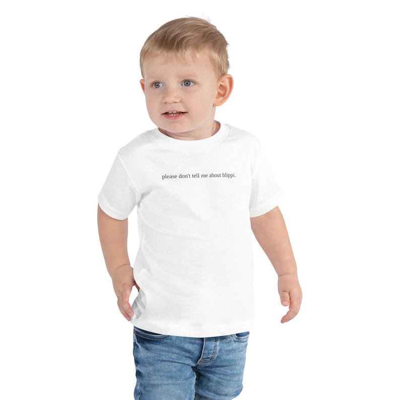 May include: A white toddler t-shirt with the text "please don't tell me about blippi." The shirt is worn by a young child wearing blue jeans. The child is smiling and looking towards the camera.