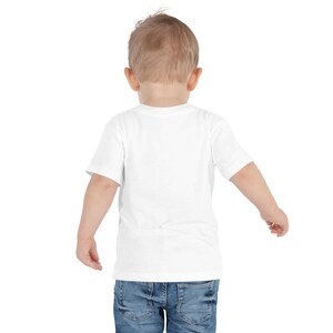 May include: A white short-sleeved t-shirt and blue denim jeans on a toddler. The child is facing away from the camera, with arms slightly outstretched. The background is plain white.