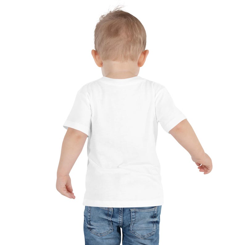 May include: A white short-sleeved t-shirt and blue denim jeans on a toddler. The child is facing away from the camera, with arms slightly outstretched. The background is plain white.